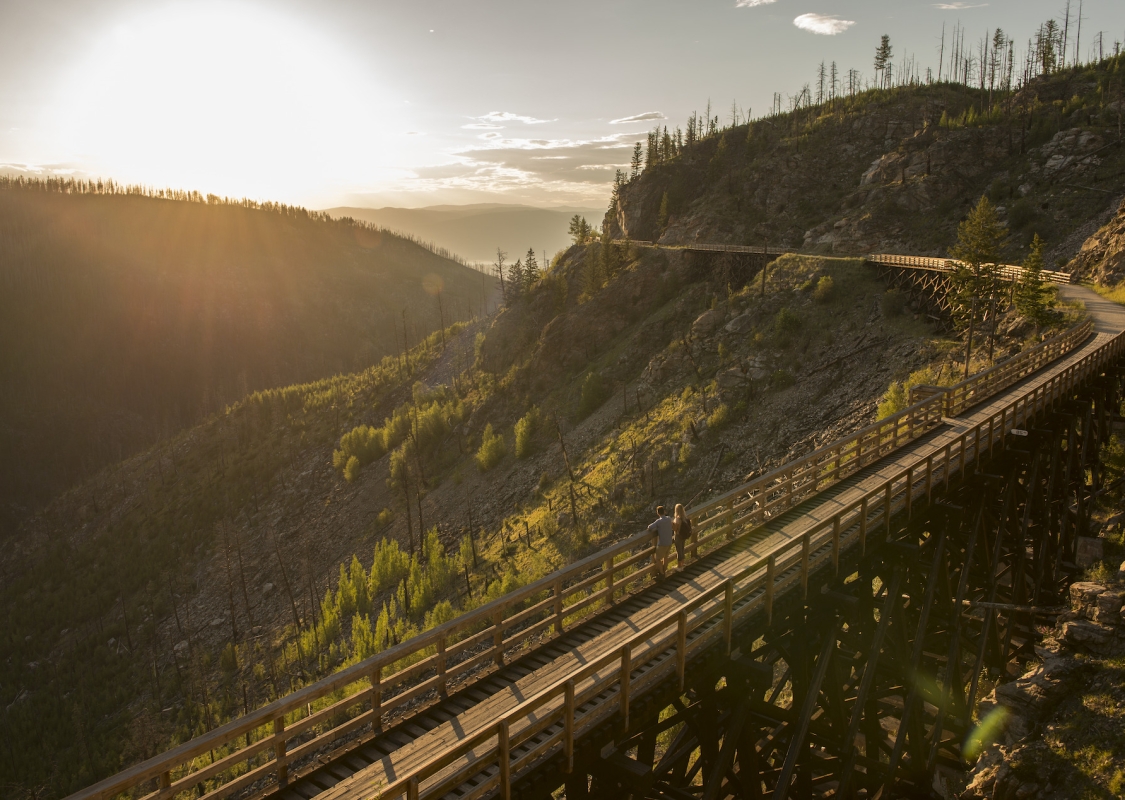 Ein Paar steht auf einer Brücke und blickt auf einen Abschnitt des Kettle Valley Rail Trail. Die Sonne geht unter. Der Rail Trail schlängelt sich hinter ihnen um den Berg.