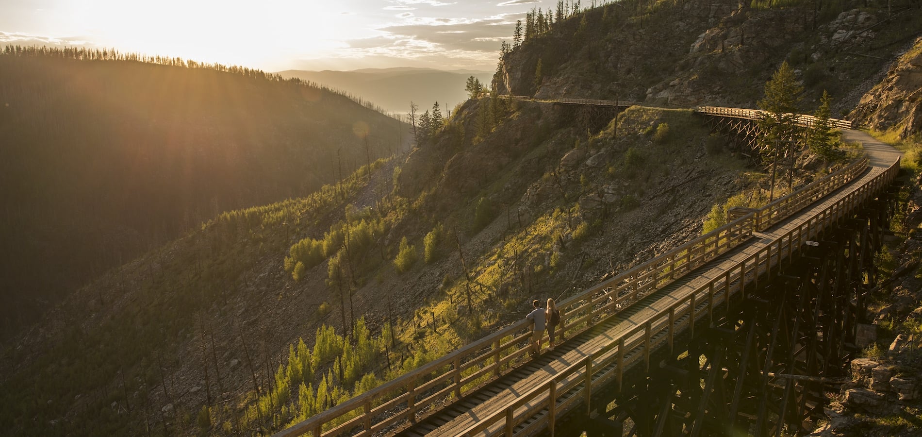 Ein Paar steht auf einer Brücke und blickt auf einen Abschnitt des Kettle Valley Rail Trail. Die Sonne geht unter. Der Rail Trail schlängelt sich hinter ihnen um den Berg.