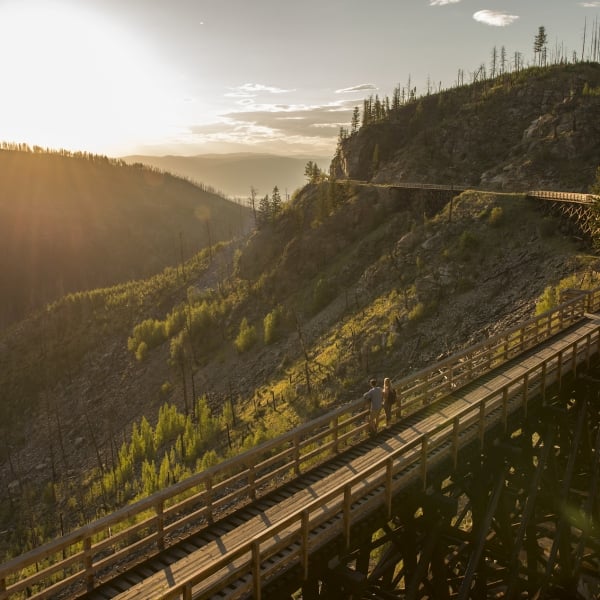 A couple stands on a bridge looking down on a section of the Kettle Valley Rail Trail. The sun is setting. The rail trail wraps around the mountain behind them.