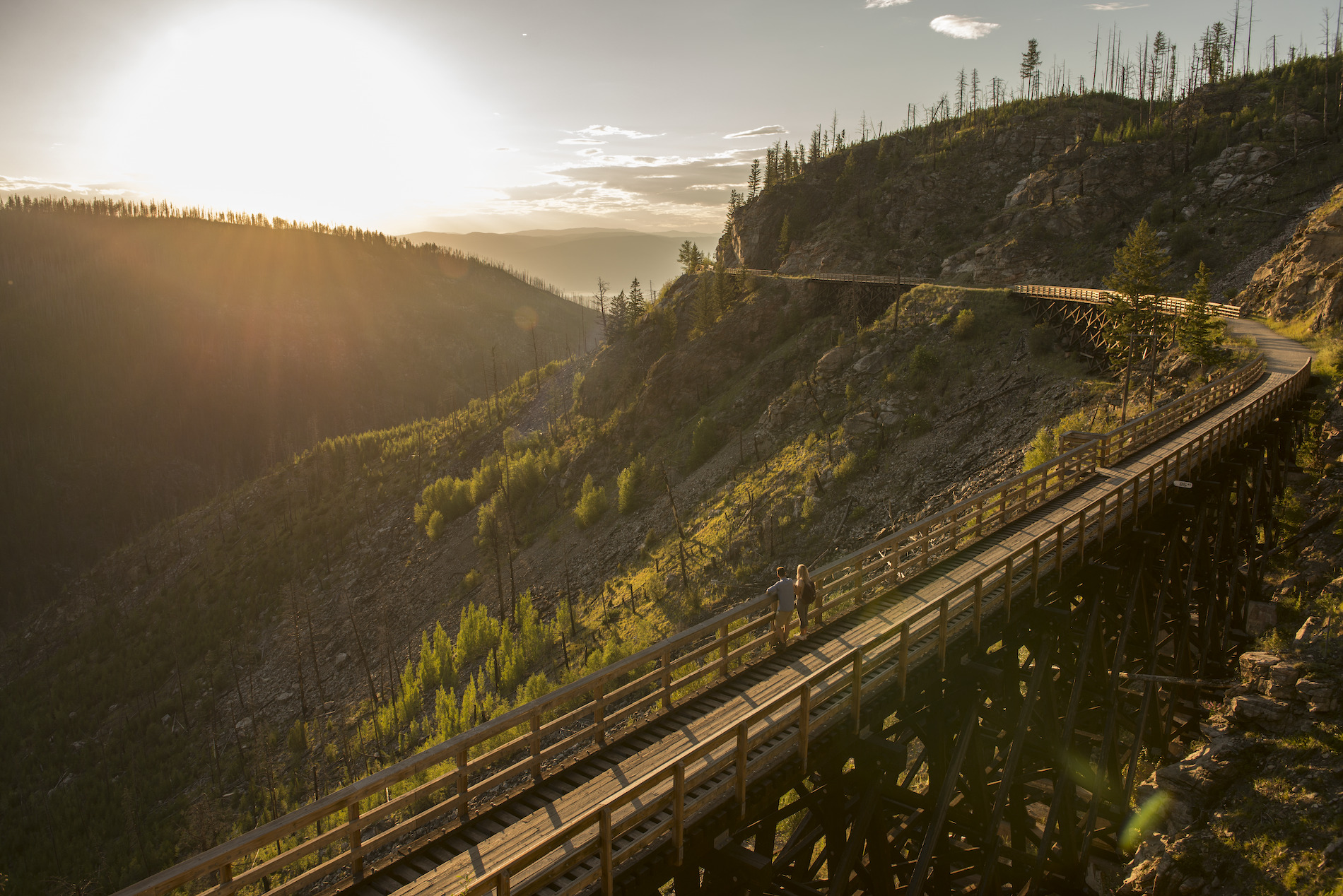 A couple stands on a bridge looking down on a section of the Kettle Valley Rail Trail. The sun is setting. The rail trail wraps around the mountain behind them.