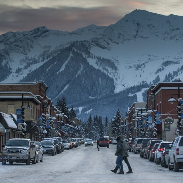 People walk through downtown Fernie in the snow. Snowcapped mountains frame the town in the distance.