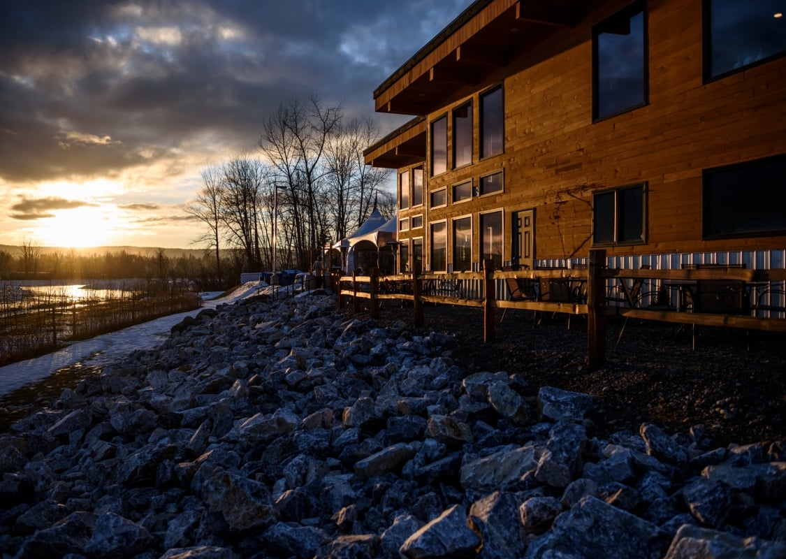 A wooden building sits on top of a rocky shore beside a river as the sun sets.