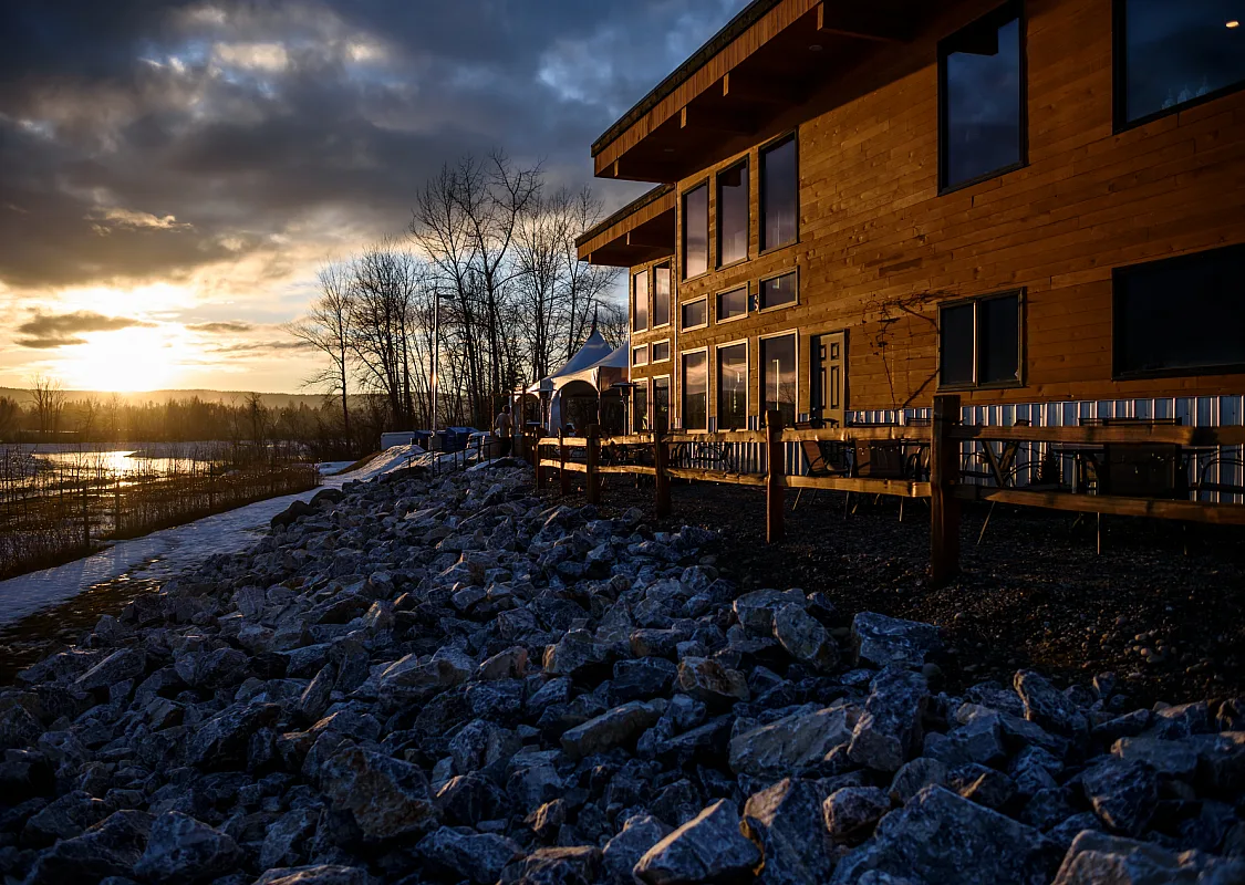 A wooden building sits on top of a rocky shore beside a river as the sun sets.
