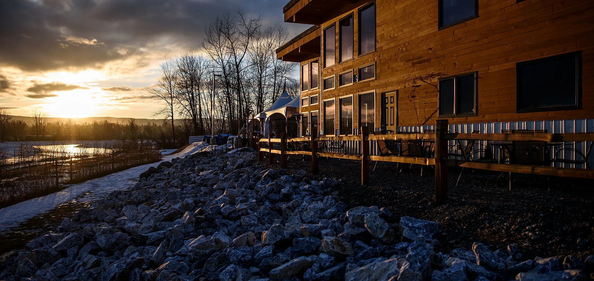 A wooden building sits on top of a rocky shore beside a river as the sun sets.