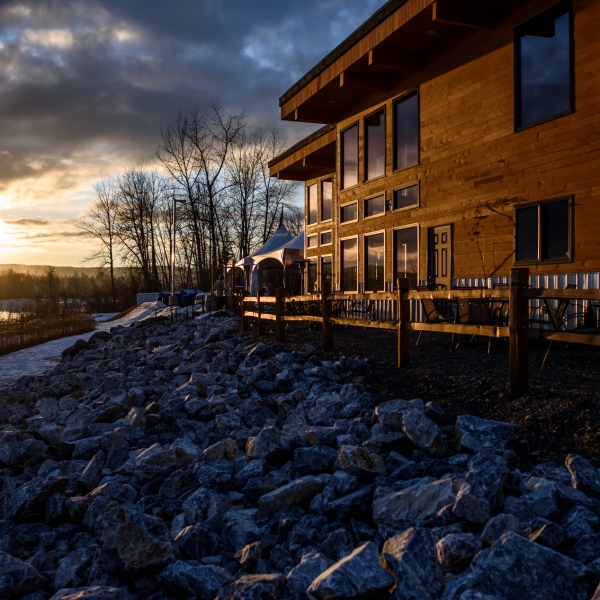 A wooden building sits on top of a rocky shore beside a river as the sun sets.