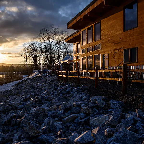 A wooden building sits on top of a rocky shore beside a river as the sun sets.