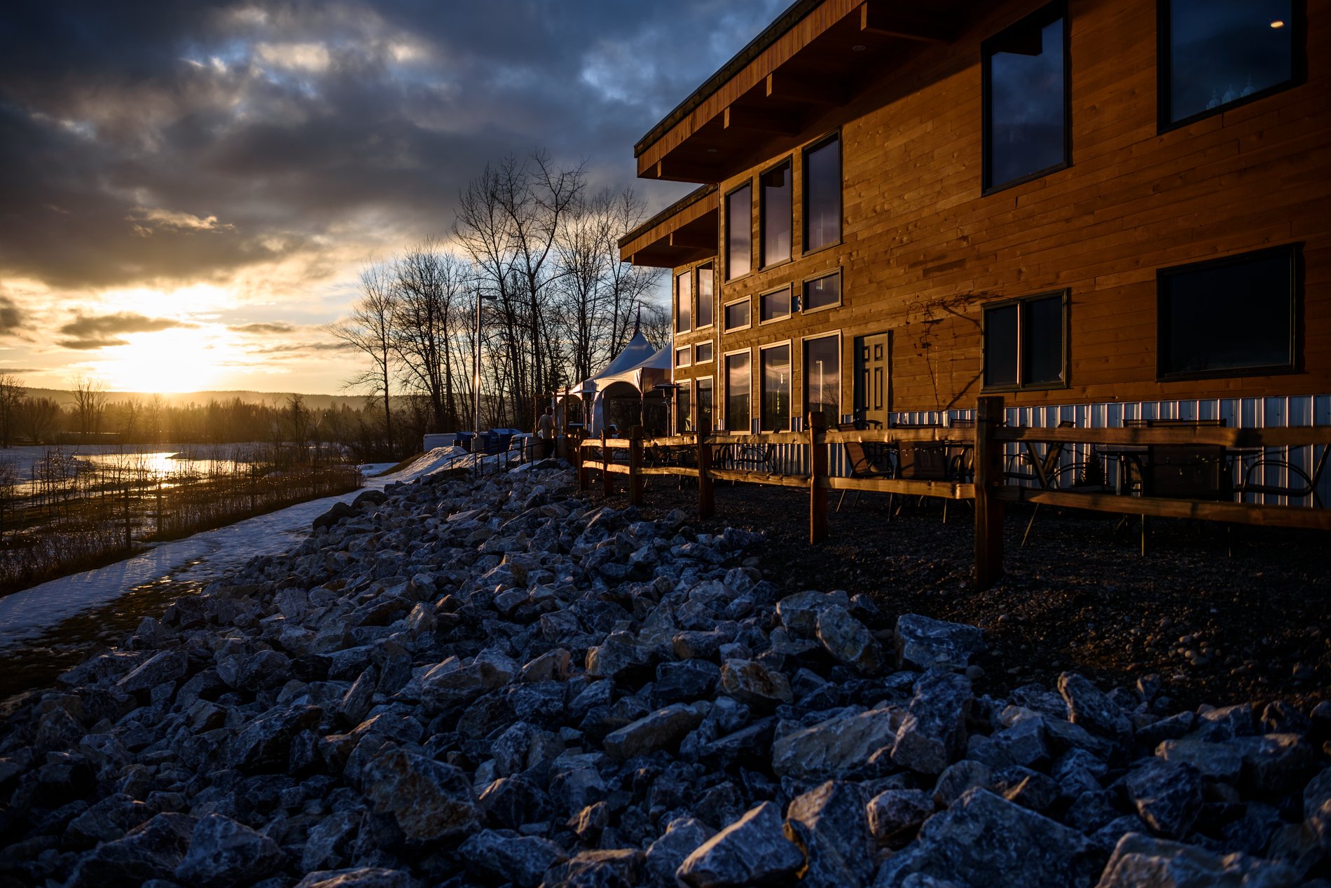 A wooden building sits on top of a rocky shore beside a river as the sun sets.