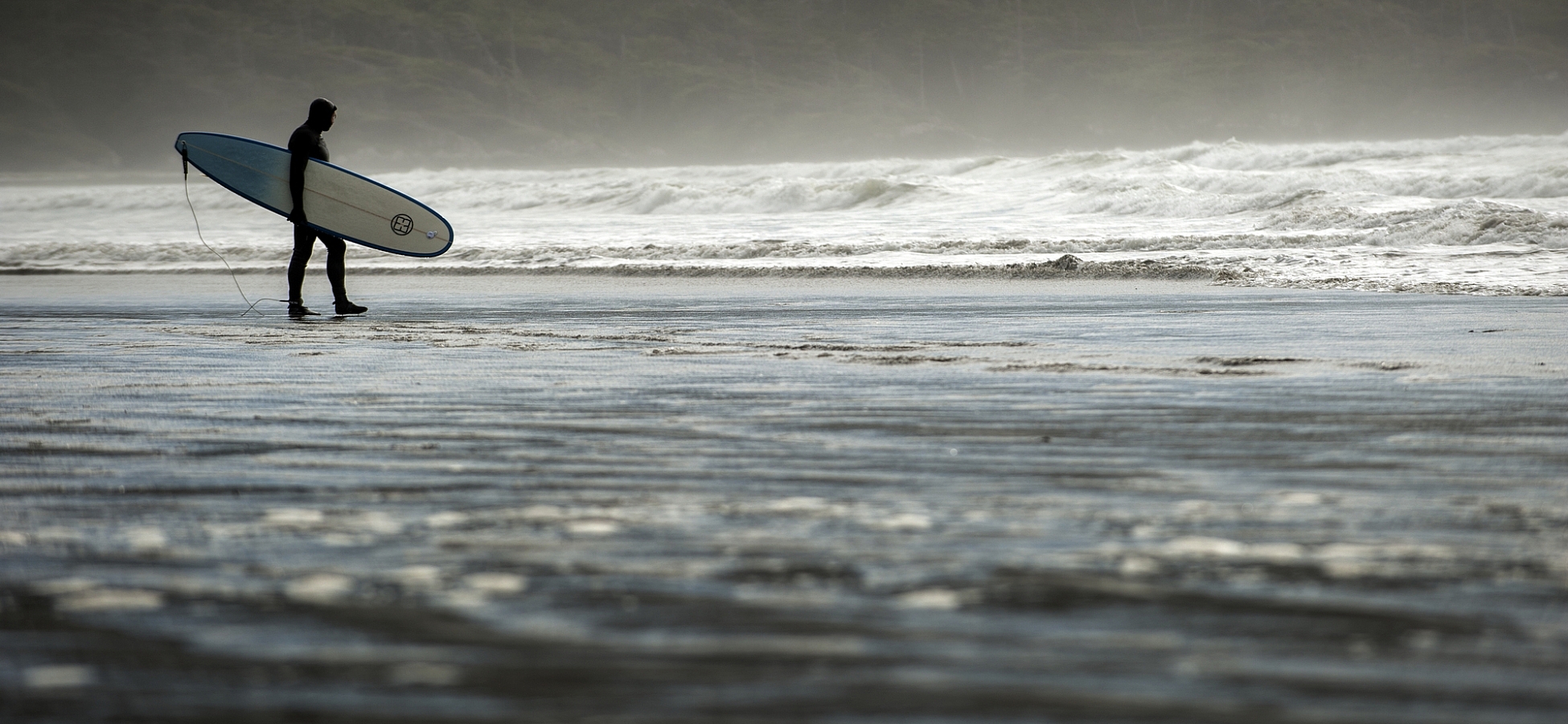 A person holds a surfboard and looks out at the water at Cox Bay