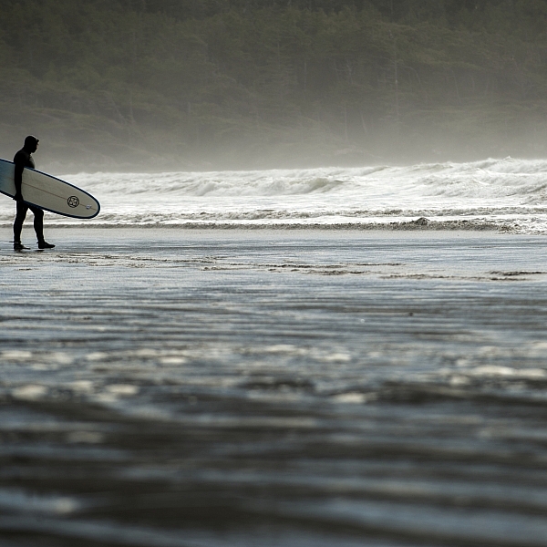 Eine Person hält ein Surfbrett und blickt in der Cox Bay auf das Wasser.