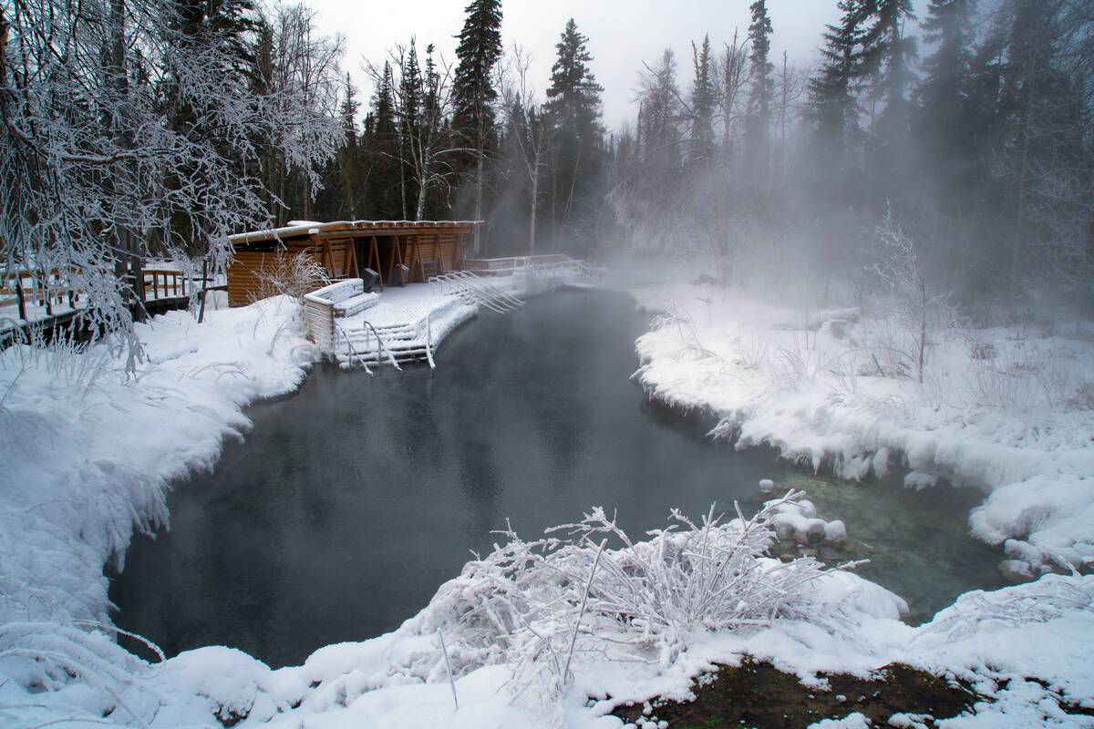 Steam rises from a small body of water surrounded by snow with a wooden structure in the background.