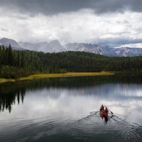 People paddle in a canoe across a lake surrounded by dense forest with mountains in the backdrop.