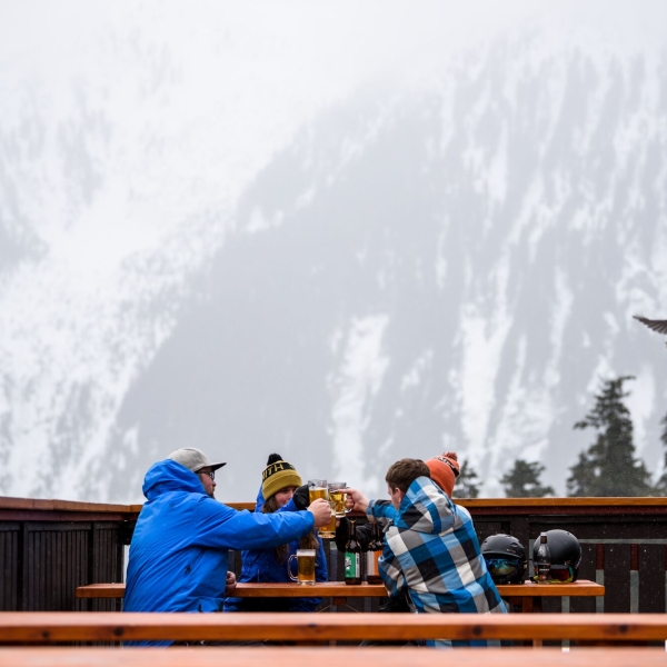 Four people in ski attire sit at a wooden picnic table on an outdoor patio with a snow-covered mountain behind them slightly obscured by fog. The four are clinking beer glasses.