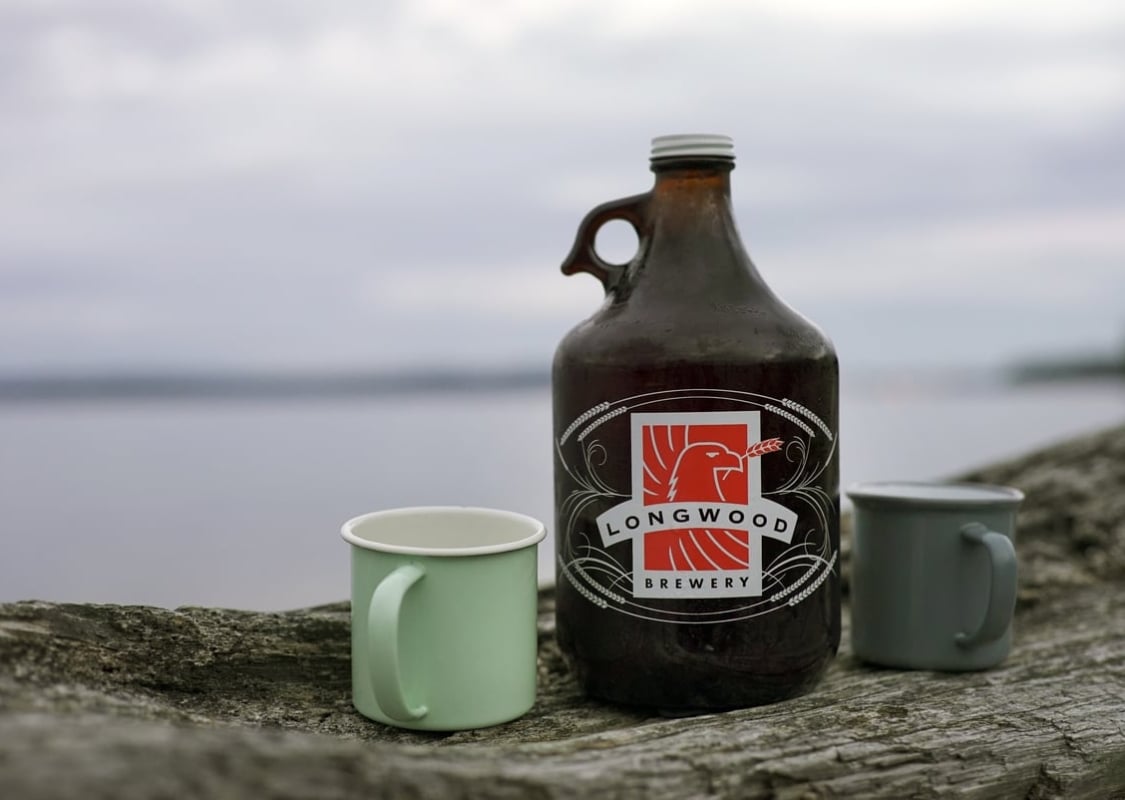 Un growler de Longwood Brewery y una taza verde descansan sobre un tronco en la playa con el océano detrás.