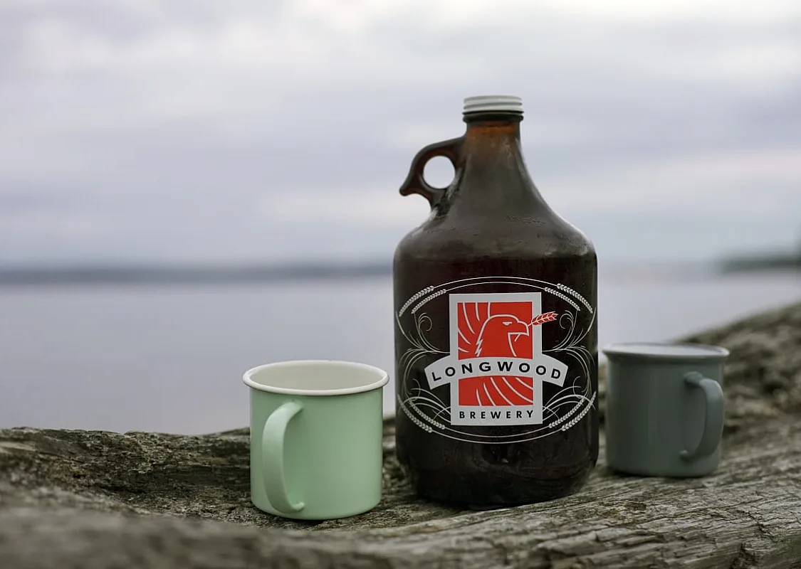 Ein Growler von Longwood Brewery und ein grüner Becher stehen auf einem Baumstamm am Strand, mit dem Ozean im Hintergrund.