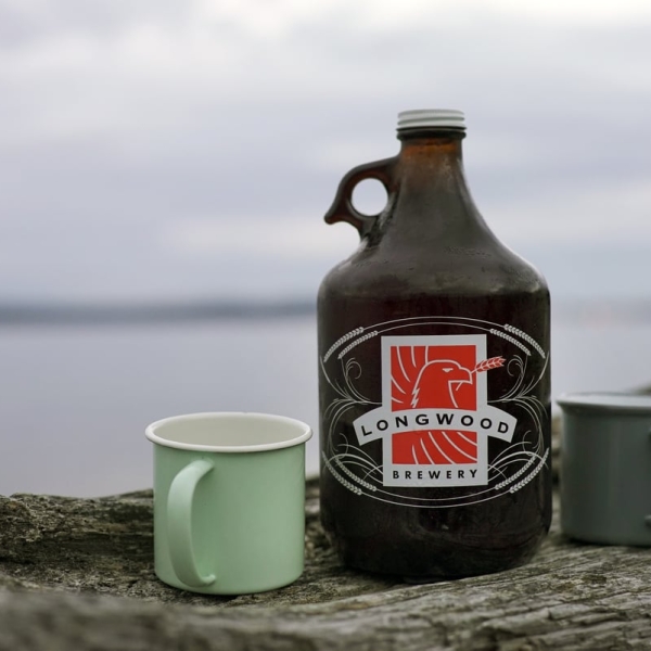 Ein Growler von Longwood Brewery und ein grüner Becher stehen auf einem Baumstamm am Strand, mit dem Ozean im Hintergrund.