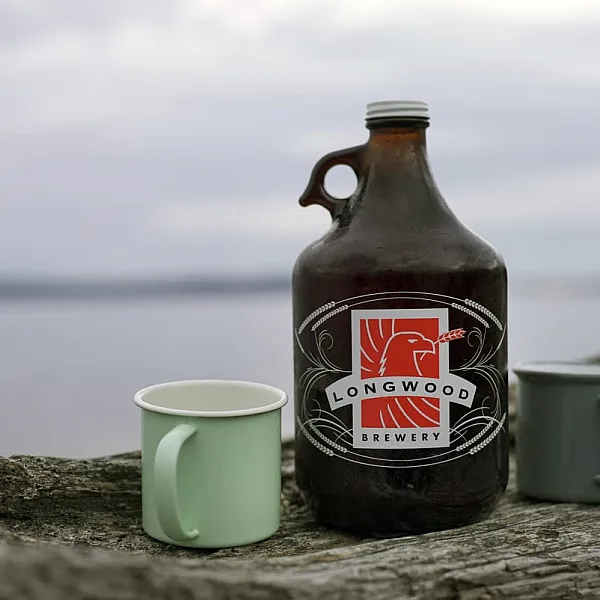 Ein Growler von Longwood Brewery und ein grüner Becher stehen auf einem Baumstamm am Strand, mit dem Ozean im Hintergrund.