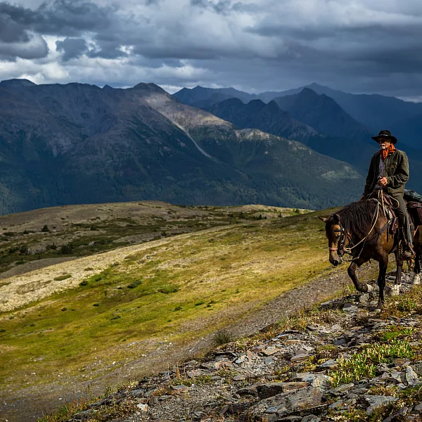 A lone horseback rider traverses the rugged countryside with rocks, sparse foliage and mountains as far as the eye can see.