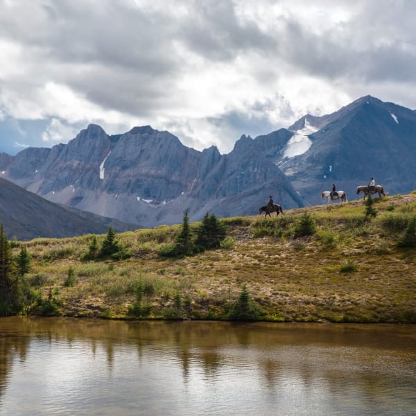 Jinetes cabalgan por una cresta con imponentes montañas de fondo, que los hacen sentir muy pequeños en comparación.