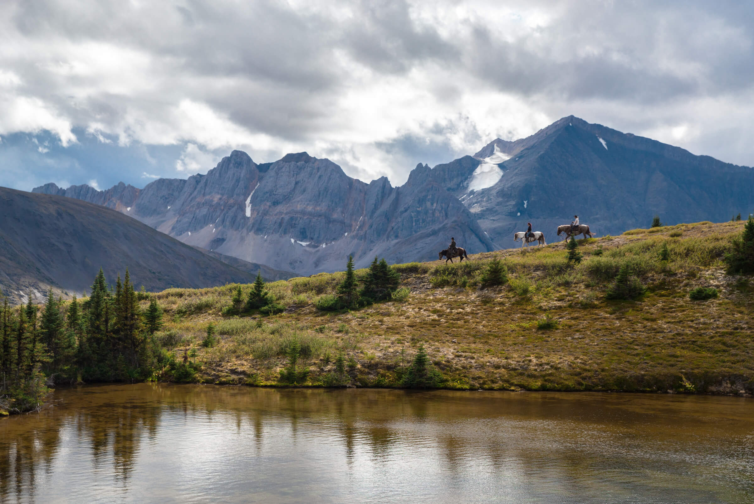 Horseback riders trot along a ridgeline with towering mountains in the backdrop, making them feel tiny in comparison.