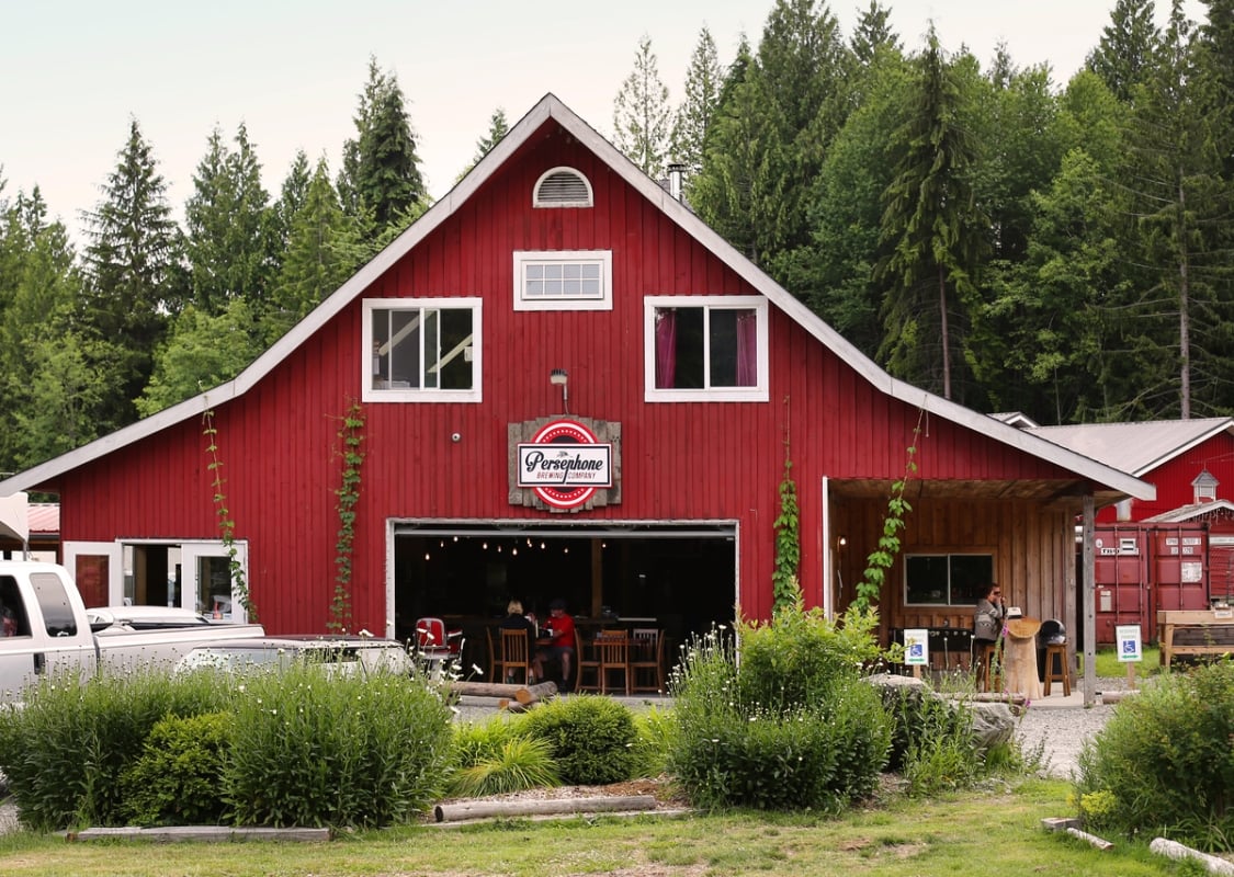 Outside the back of Persephone Brewing Company in Gibsons. The building is built like a large red barn with white trim. Tables are seen inside and cars and grass are out front.