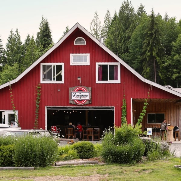 Outside the back of Persephone Brewing Company in Gibsons. The building is built like a large red barn with white trim. Tables are seen inside and cars and grass are out front.