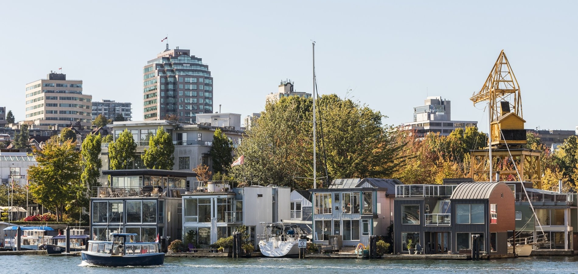 A view of a waterfront neighborhood at False Creek features modern houseboats and small buildings along the water’s edge, with taller buildings and trees in the background. A small boat cruises through the calm water in the foreground, adding a sense of activity to the scene. An old yellow crane structure stands prominently among the buildings, blending the industrial past with the vibrant, contemporary setting. The sky is clear, and the overall atmosphere is lively and picturesque, showcasing a unique blend of urban and maritime elements.