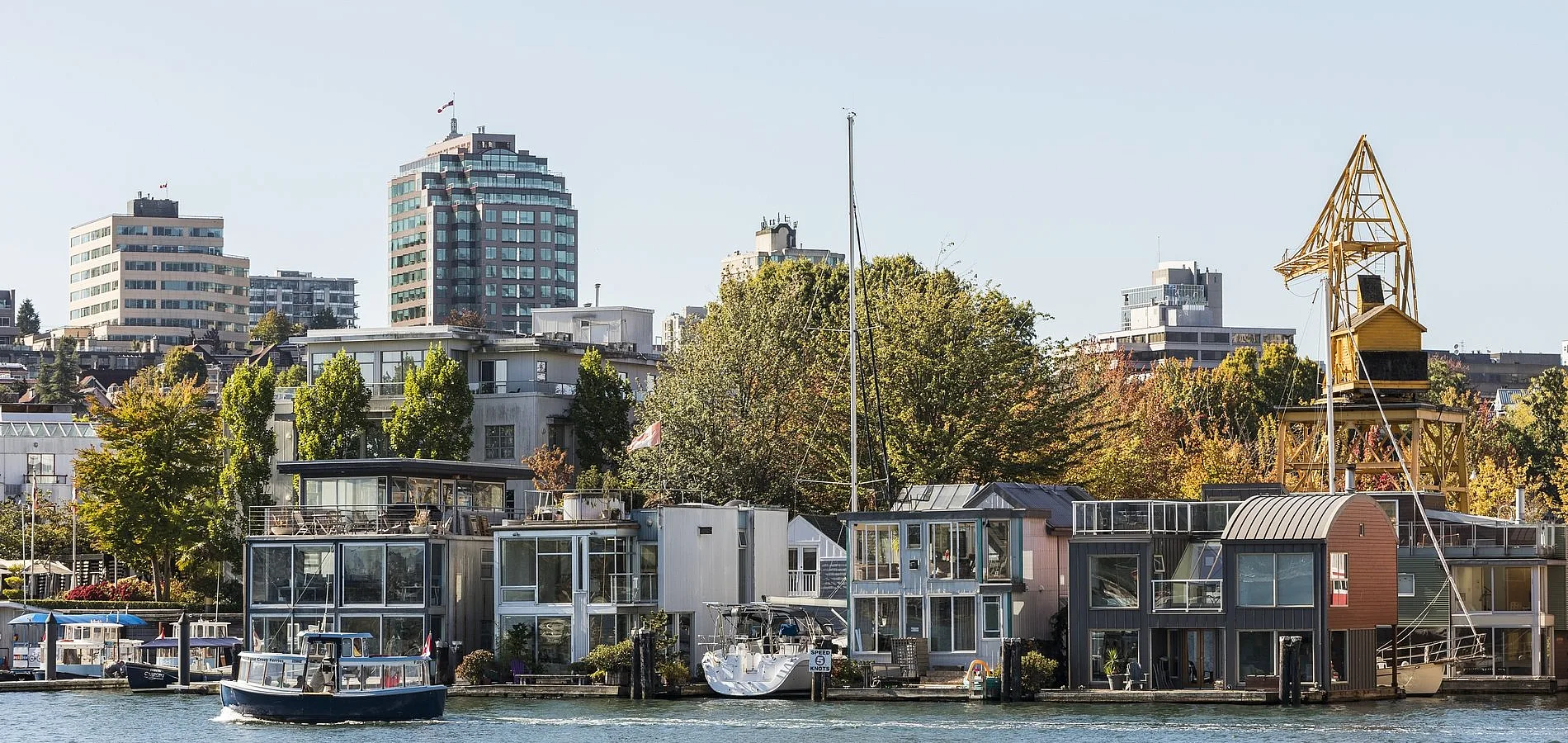 Una vista de un barrio frente al mar en False Creek muestra modernas casas flotantes y pequeños edificios a lo largo de la orilla, con edificios más altos y árboles al fondo. Un pequeño bote navega por las tranquilas aguas en primer plano, añadiendo una sensación de actividad a la escena. Una antigua estructura de grúa amarilla se alza prominentemente entre los edificios, mezclando el pasado industrial con el vibrante entorno contemporáneo. El cielo está despejado y el ambiente general es animado y pintoresco, mostrando una mezcla única de elementos urbanos y marítimos.