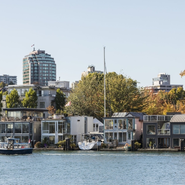 Eine Ansicht eines Viertels am Wasser am False Creek zeigt moderne Hausboote und kleine Gebäude entlang des Wassers, mit höheren Gebäuden und Bäumen im Hintergrund. Ein kleines Boot fährt durch das ruhige Wasser im Vordergrund, was der Szene Lebendigkeit verleiht. Eine alte gelbe Kranstruktur ragt prominent zwischen den Gebäuden hervor und verbindet die industrielle Vergangenheit mit der lebendigen, zeitgenössischen Umgebung. Der Himmel ist klar und die Gesamtatmosphäre ist lebhaft und malerisch, was eine einzigartige Mischung aus urbanen und maritimen Elementen zeigt.