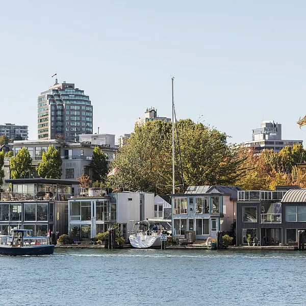 Una vista de un barrio frente al mar en False Creek muestra modernas casas flotantes y pequeños edificios a lo largo de la orilla, con edificios más altos y árboles al fondo. Un pequeño bote navega por las tranquilas aguas en primer plano, añadiendo una sensación de actividad a la escena. Una antigua estructura de grúa amarilla se alza prominentemente entre los edificios, mezclando el pasado industrial con el vibrante entorno contemporáneo. El cielo está despejado y el ambiente general es animado y pintoresco, mostrando una mezcla única de elementos urbanos y marítimos.