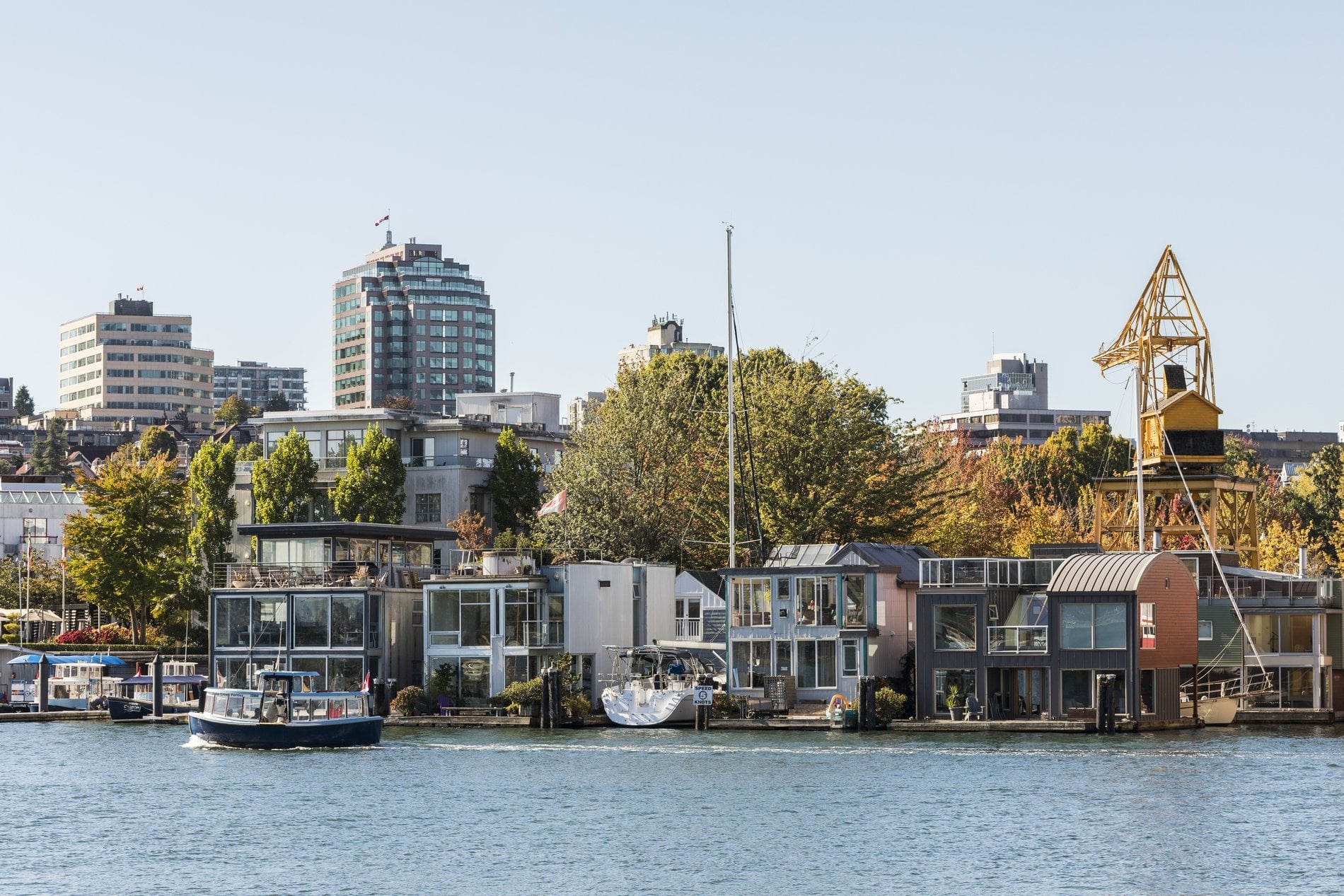 Una vista del barrio costero de False Creek muestra modernas casas flotantes y pequeños edificios a lo largo de la orilla, con edificios más altos y árboles al fondo. Una pequeña embarcación navega por las tranquilas aguas en primer plano, añadiendo una sensación de actividad a la escena. Una vieja grúa amarilla destaca entre los edificios, mezclando el pasado industrial con el vibrante entorno contemporáneo. El cielo está despejado y el ambiente general es animado y pintoresco, mostrando una mezcla única de elementos urbanos y marítimos.