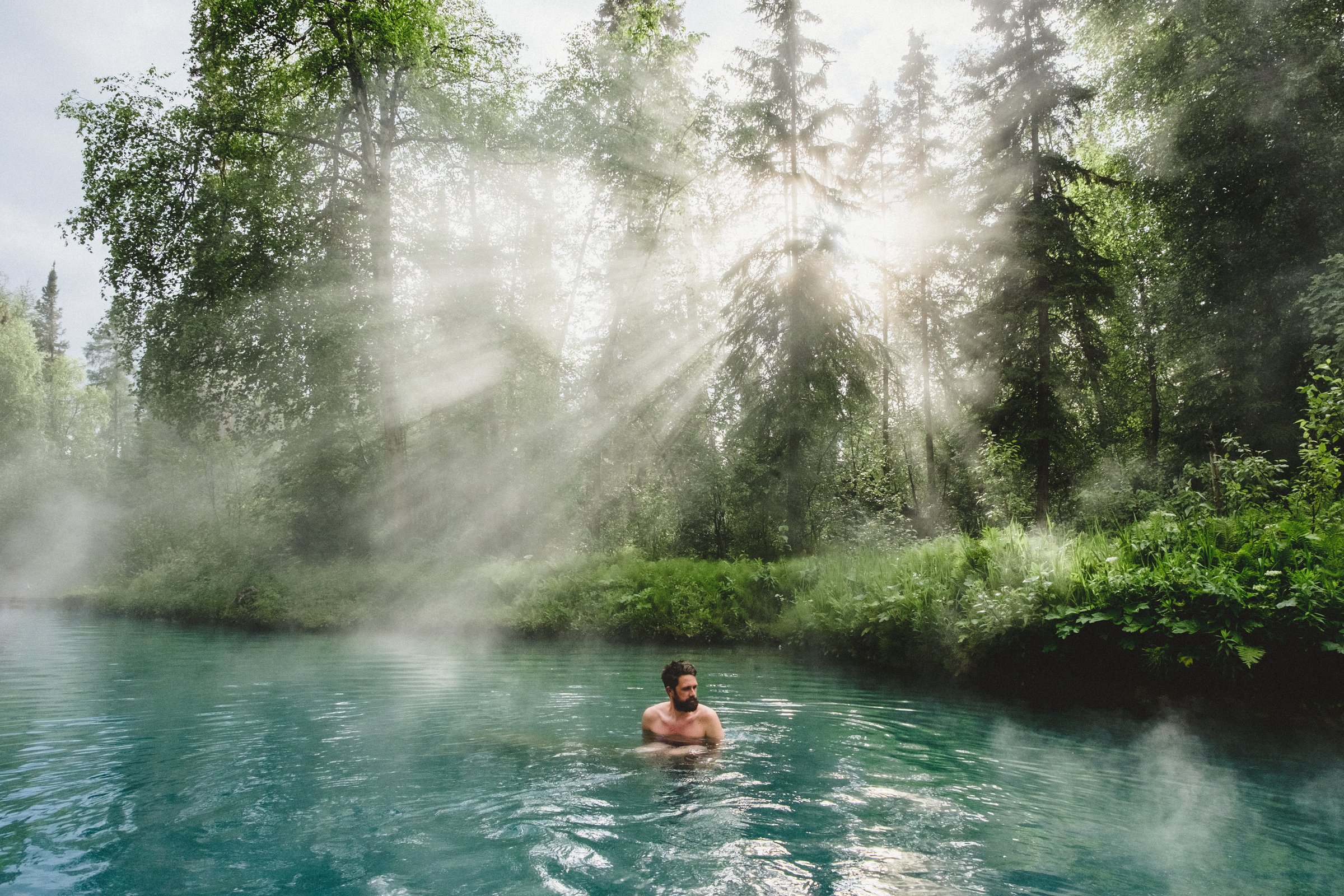 A person soaks in Liard hot springs with light rays peeking through the trees.