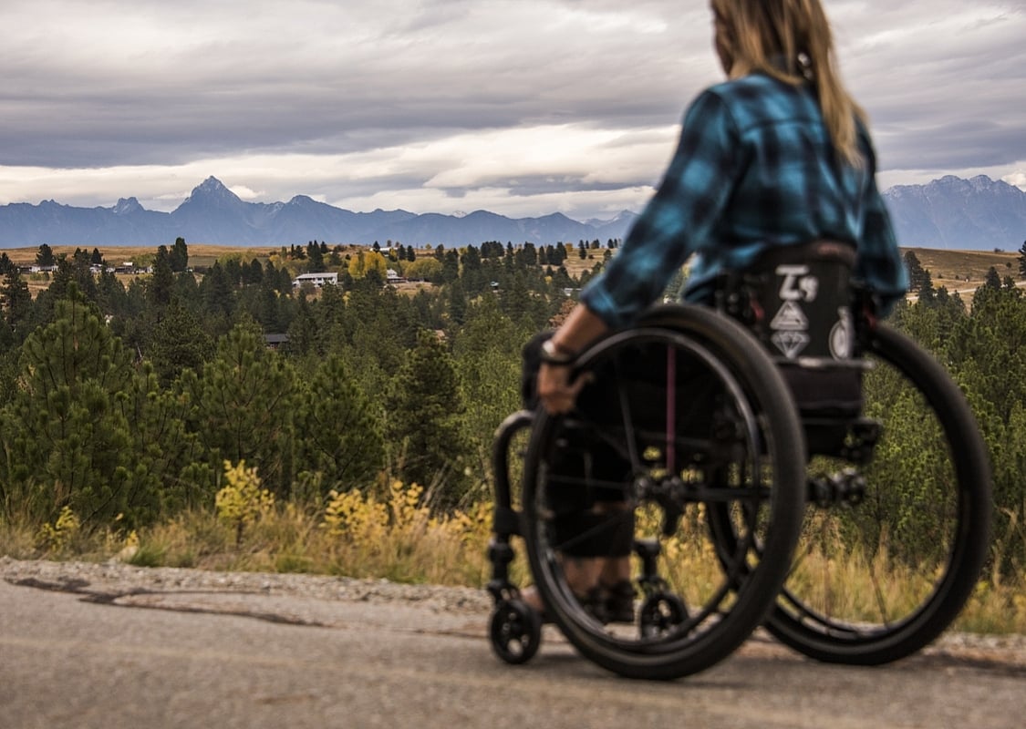 A wheelchair user wearing a blue and black plaid shirt, is on a trail, looking out into the distance. There are evergreen trees and a mountain in the background.