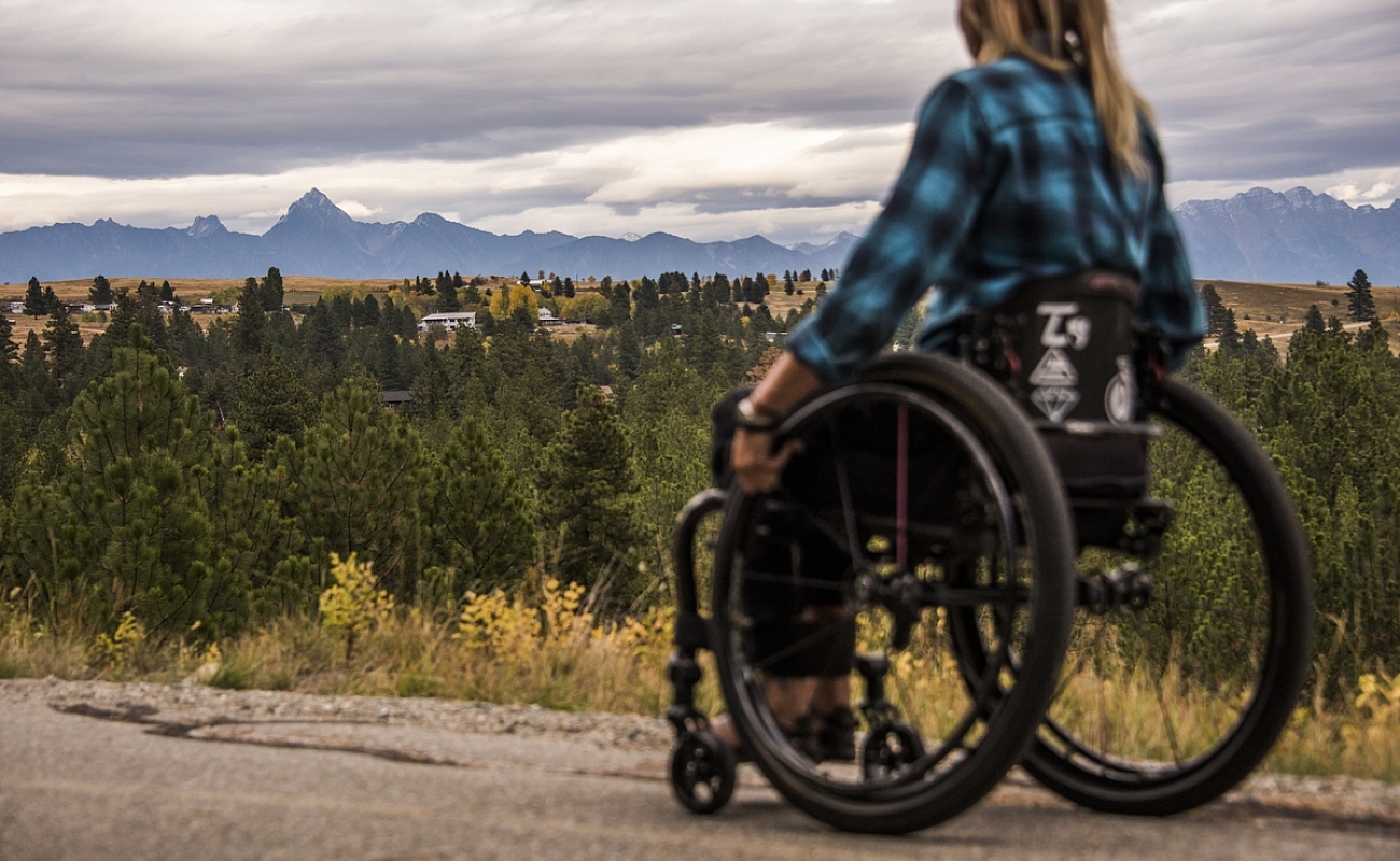 A wheelchair user wearing a blue and black plaid shirt, is on a trail, looking out into the distance. There are evergreen trees and a mountain in the background.