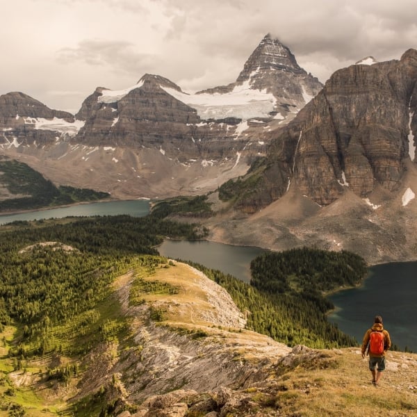 A hiker stands on the right of the frame, small, on a ledge overlooking a clear mountain lake with rugged peaks rising in the distance