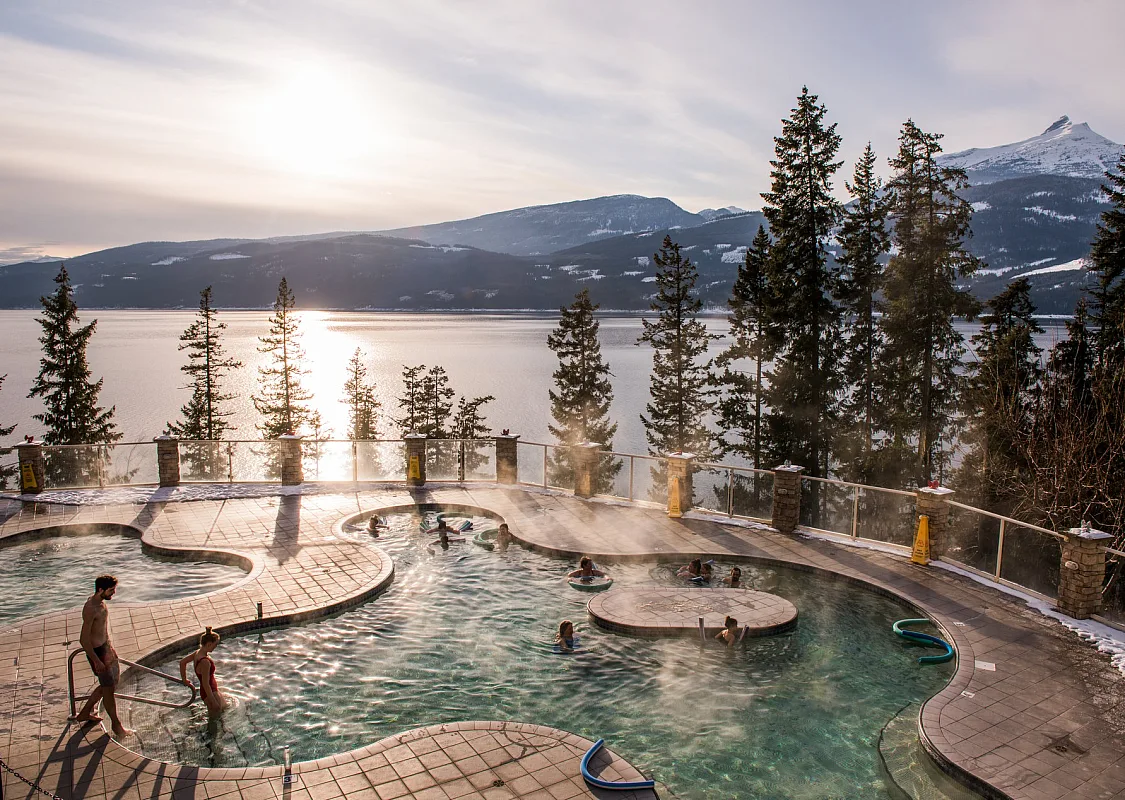 An amoeba-shaped pool with steam rising from the surface with sparse trees behind it and a large lake in the background.