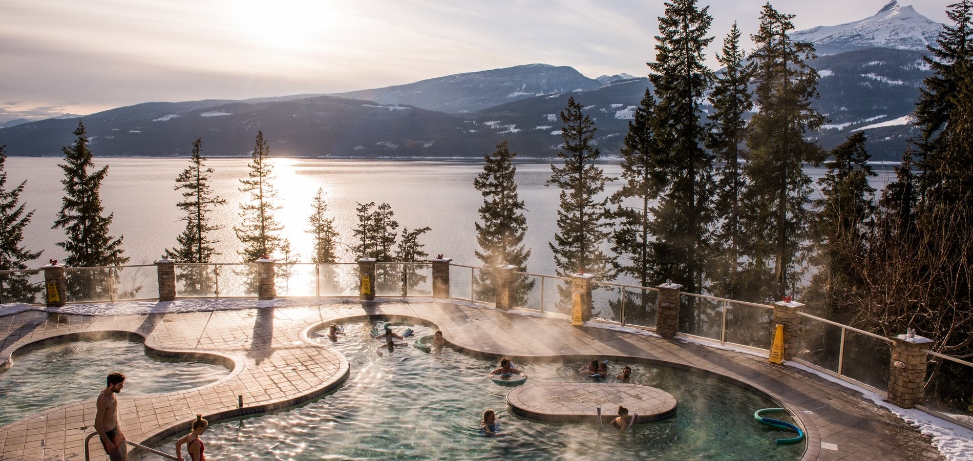 An amoeba-shaped pool with steam rising from the surface with sparse trees behind it and a large lake in the background.