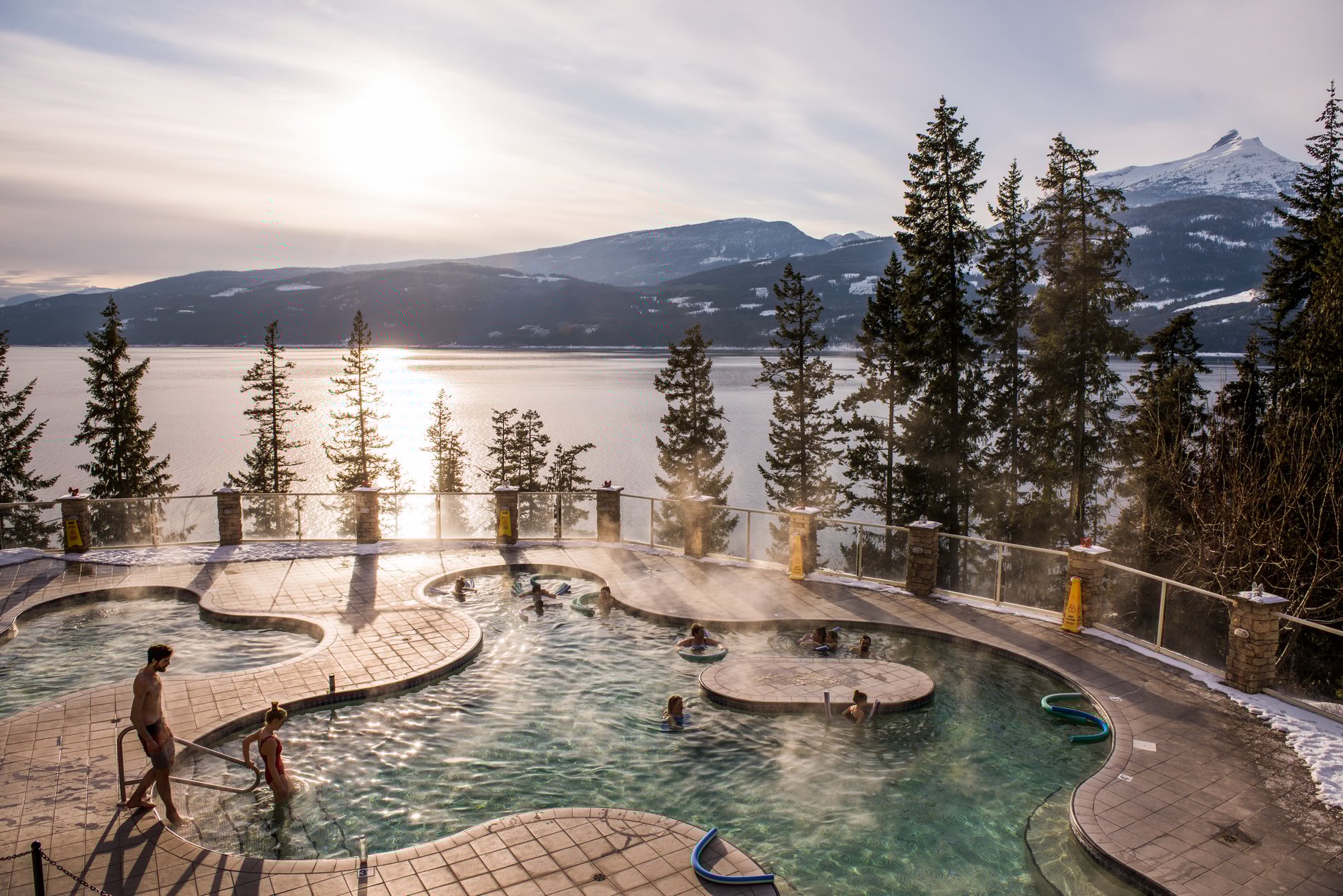 An amoeba-shaped pool with steam rising from the surface with sparse trees behind it and a large lake in the background.