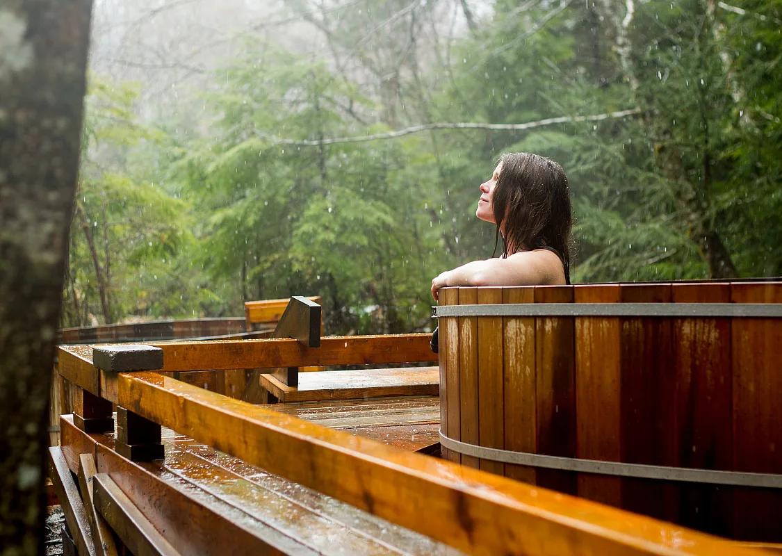 A woman soaks in a wooden tub looking out at the forest.
