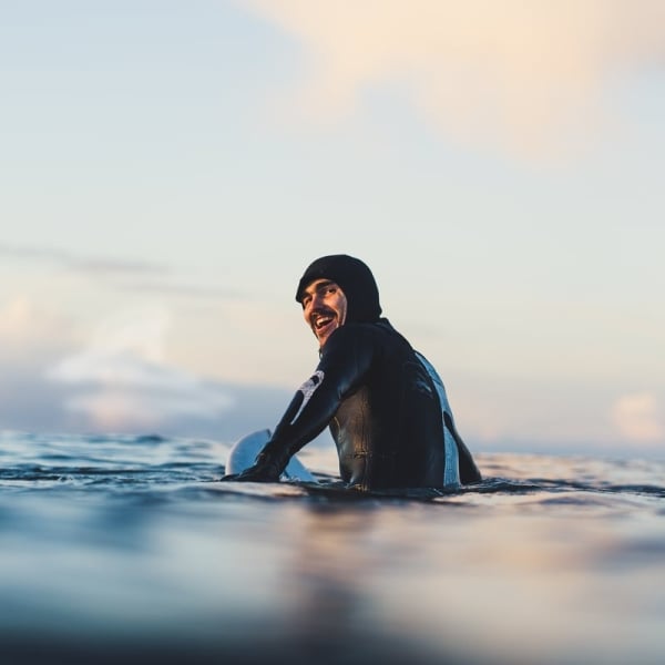 A surfer in a black wetsuit looks back at the camera from the water.