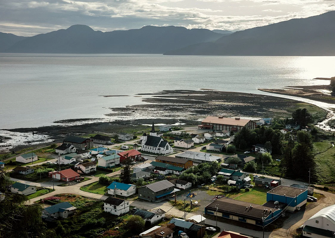 An aerial of a coastal village tucked in an inlet on the edge of the Pacific Ocean with towering mountains in the background.