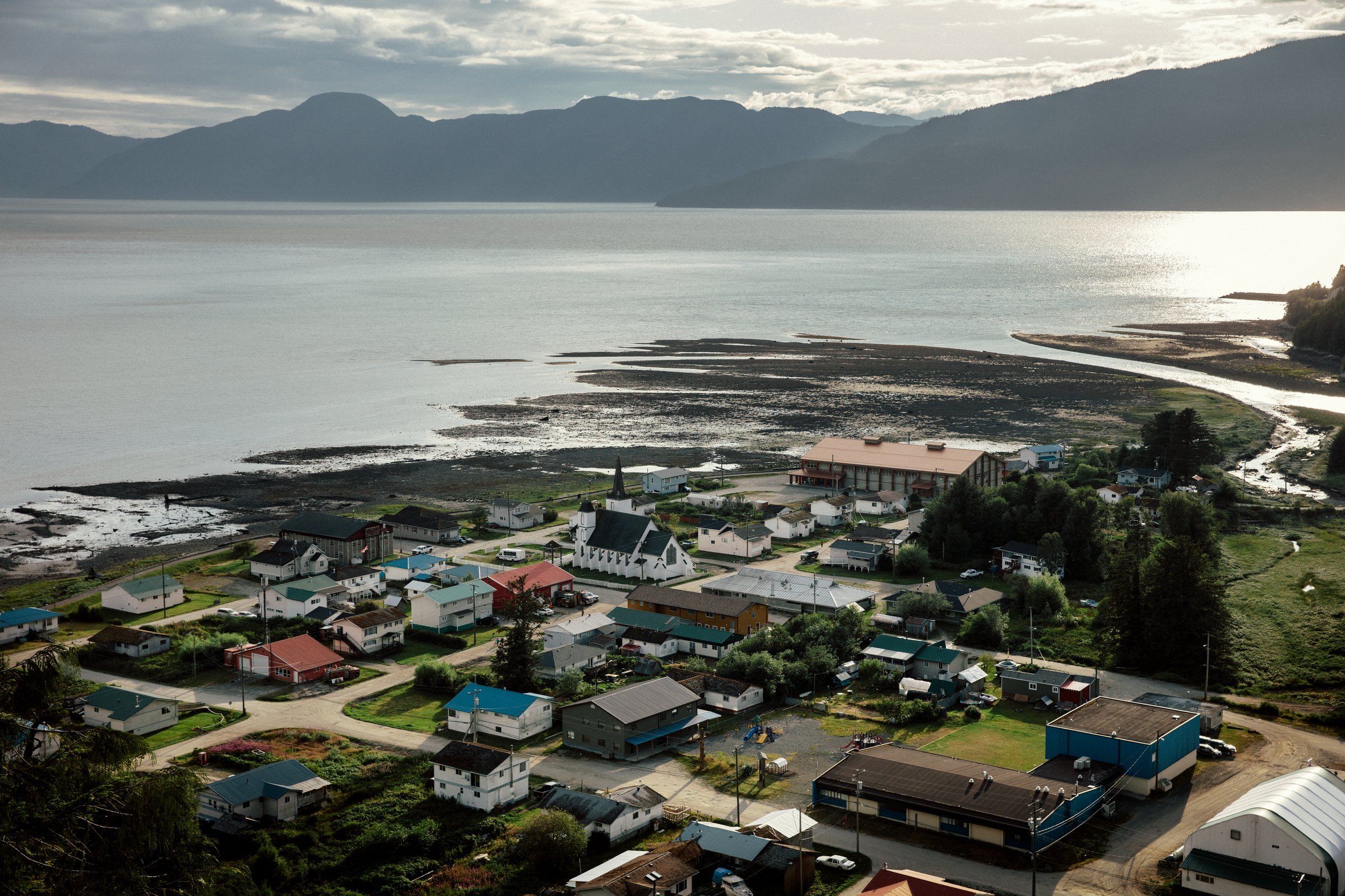 An aerial of a coastal village tucked in an inlet on the edge of the Pacific Ocean with towering mountains in the background.