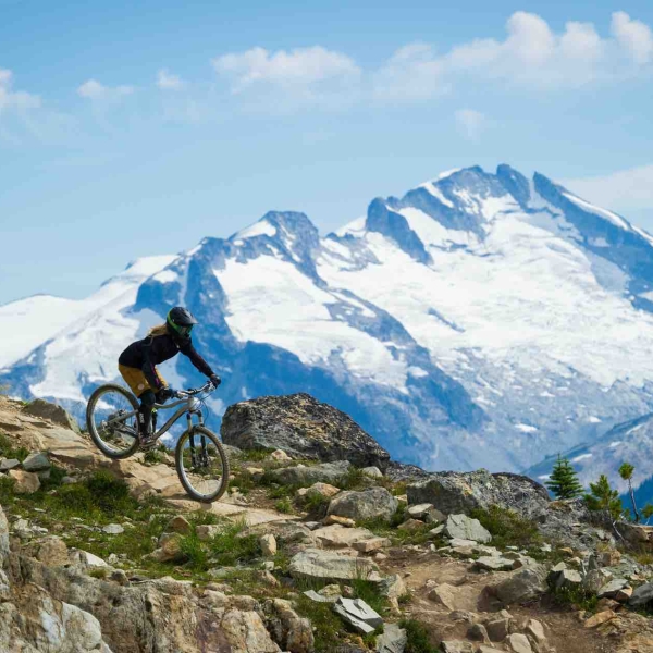 A person mountain bikes in the alpine down a rocky dirt path on the Whistler Mountain Bike Park. A snow covered mountain is in the background.