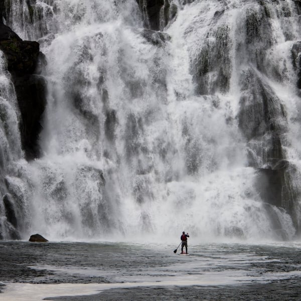 Man in the distance paddleboarding towards a cascading waterfall