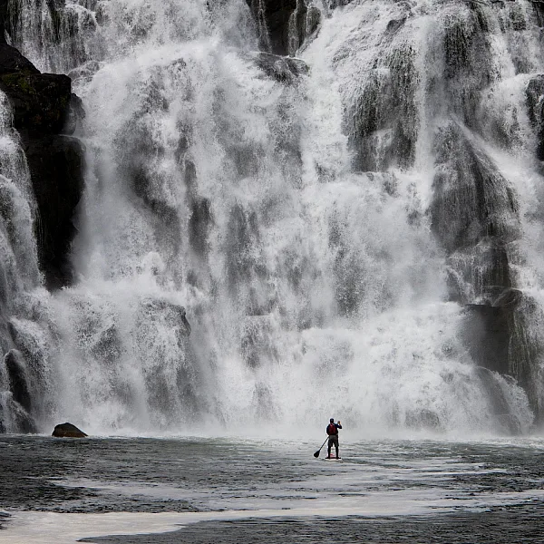 Man in the distance paddleboarding towards a cascading waterfall
