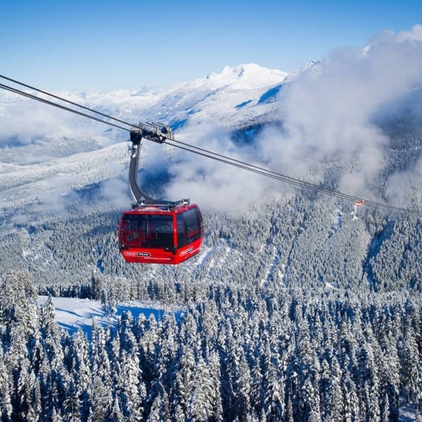 A red gondola car hangs over a snowy mountain valley with a cloudy sky and mountains behind the clouds