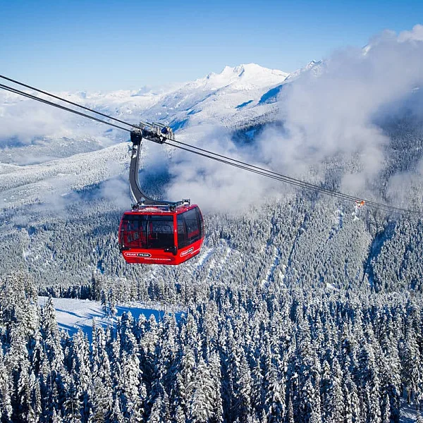 A red gondola car hangs over a snowy mountain valley with a cloudy sky and mountains behind the clouds