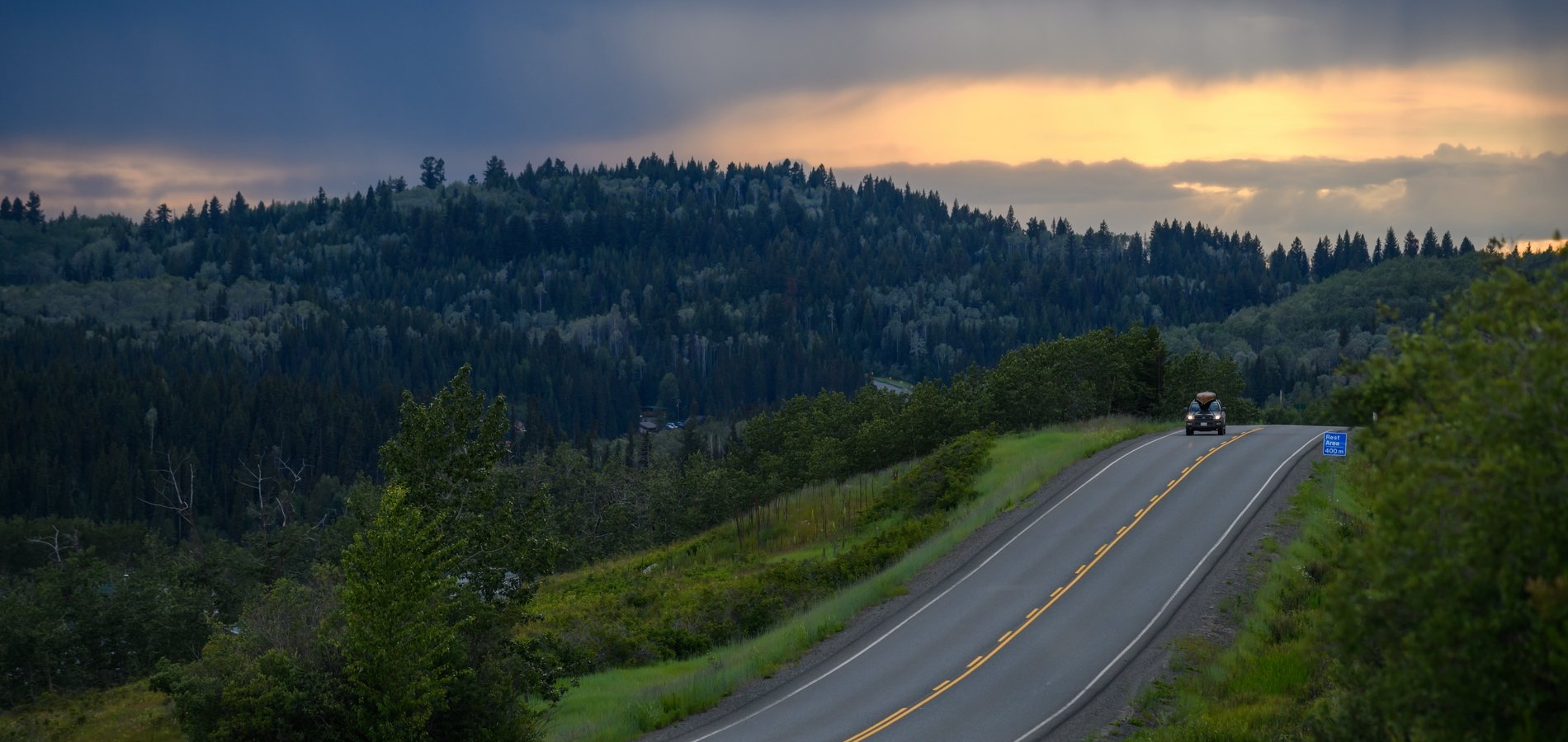 A single car drives down Highway 24 as the sun sets in the distance. The road is framed by trees on both sides. The vehicle has a canoe strapped to the roof.