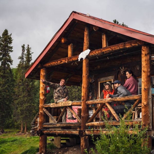 A group of people sits in a small log shelter fishing camp, surrounded by wilderness.