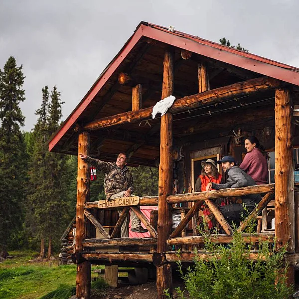 Un grupo de personas se sienta en un pequeño campamento de pesca con refugio de troncos, rodeado de naturaleza salvaje.