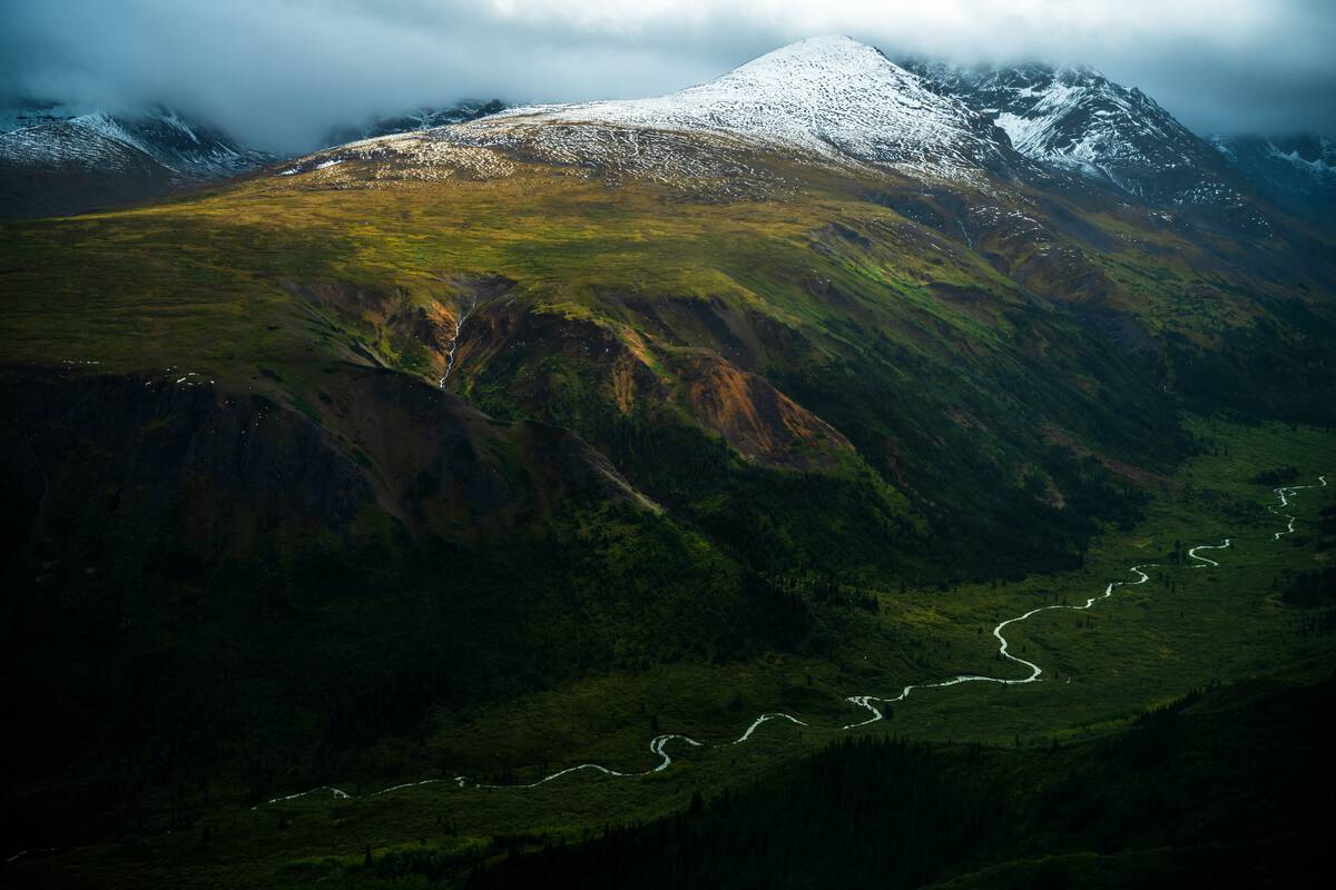 A bird's eye view of a lush river valley surrounded by a patchwork of colourful snow-capped mountains.